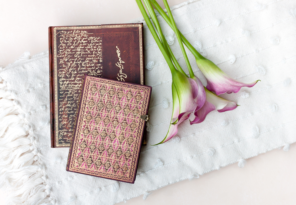 Paperblanks Shakespeare, Sir Thomas More and River Cascade Alluvium journals with pink flowers on a white blanket.